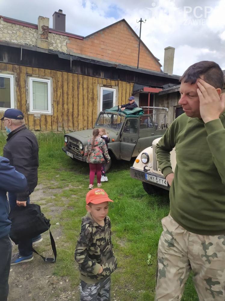 Zdjęcie: Grupa mężczyzn i dzieci stoi, a w tle drewniany budynek i wojskowe samochody terenowe